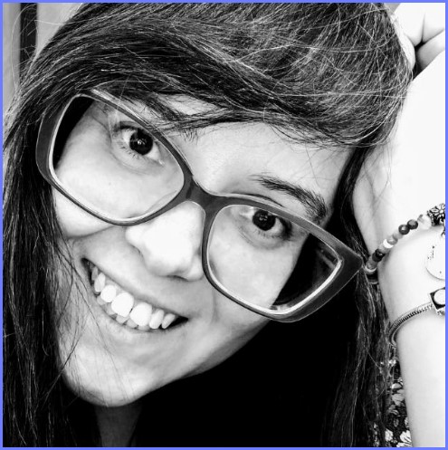 Foto em preto e branco de Ana Cândida, ela usa óculos quadrados grandes e sorri com a mão posicionada na cabeça.