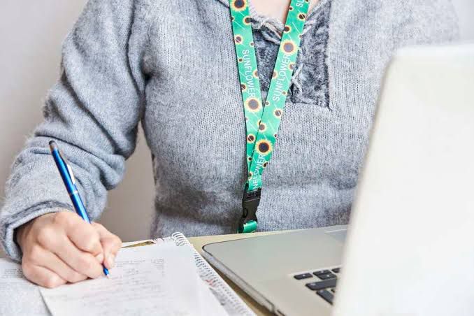 Foto de uma pessoa trabalhando ao computador. Ela usa uma blusa cinza e escreve em folhas de papel brancas que estão ao lado de seu notebook. Ela usa uma fita verde com estampa de girassol em torno do pescoço.